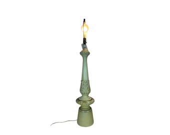 May include: A tall, sage green table lamp with a decorative, carved design. The lamp has a bulb at the top and a cord extending from the base. The lamp's design includes a detailed, carved pattern and a round base.