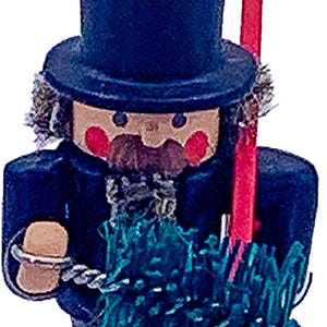 May include: A wooden figurine of a chimney sweep, wearing a dark blue top hat and coat. The figurine has a tan face with red cheeks and a brown mustache. It holds a red pole and a green brush.