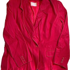 Op de afbeelding: Een felrode blazer met een enkele knoopsluiting en drie knopen op elke mouw. De blazer heeft een klassieke revers en een zak aan de voorkant. De onderkant van de blazer wordt gecombineerd met een geruite rok.