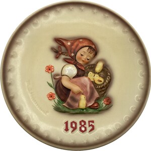Könnte beinhalten: Ein Porzellanteller mit einem Reliefbild eines Mädchens in einem rot-weißen Kleid, das einen Korb mit Küken hält. Der Teller ist mit einem Blumenmuster und dem Jahr 1985 verziert.