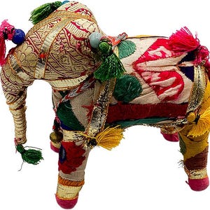 May include: A decorative elephant figurine with ornate detailing. The elephant is covered in patterned fabric with gold trim, red, green, and yellow tassels, and colorful bead accents. The trunk is curled, and the legs are short and sturdy.
