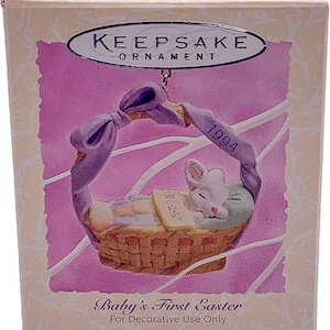May include: A white baby bunny sleeping in a wicker basket with a purple ribbon and a sign that says "Welcome". The basket is decorated with pastel colors and the year 1994 is printed on the ribbon. The box reads "Keepsake Ornament Baby's First Easter For Decorative Use Only".