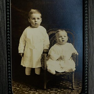 May include: Vintage sepia-toned photograph of two young children. One child stands, the other sits in a wicker chair. Both are dressed in white outfits. The background is a dark blue and brown, with a decorative border.