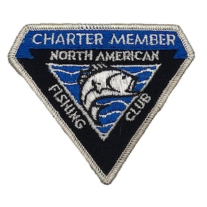 May include: A diamond-shaped patch with the text "CHARTER MEMBER NORTH AMERICAN FISHING CLUB" in white lettering. The patch features a blue and black colour scheme with a fish graphic in the centre. The border is silver.