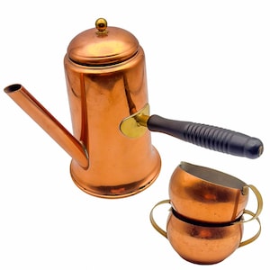 May include: A copper coffee pot with a dark wooden handle and two copper cups. The coffee pot has a long spout and a lid with a small, gold-colored knob. The cups have small handles and are stacked on top of each other.
