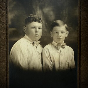 May include: Sepia-toned portrait of two young boys. Both are wearing striped button-down shirts with bow ties. The boy on the left has a rounder face and the boy on the right has a more slender face. The background is a blurred, dark landscape.