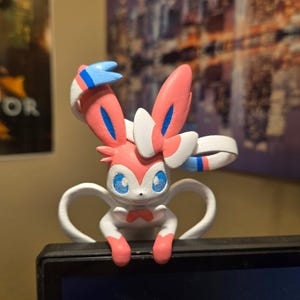 May include: A colorful figurine of a Sylveon character, featuring pink, white, and blue accents. The figure has large ears, blue eyes, and a bow tie. The figurine is perched on a black surface.