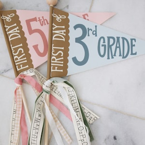 May include: Two pennant flags with wooden sticks and colorful ribbons. The first flag is pink with the text "FIRST DAY of 5th" in brown and pink. The second flag is blue with the text "FIRST DAY of 3rd GRADE" in brown and blue.