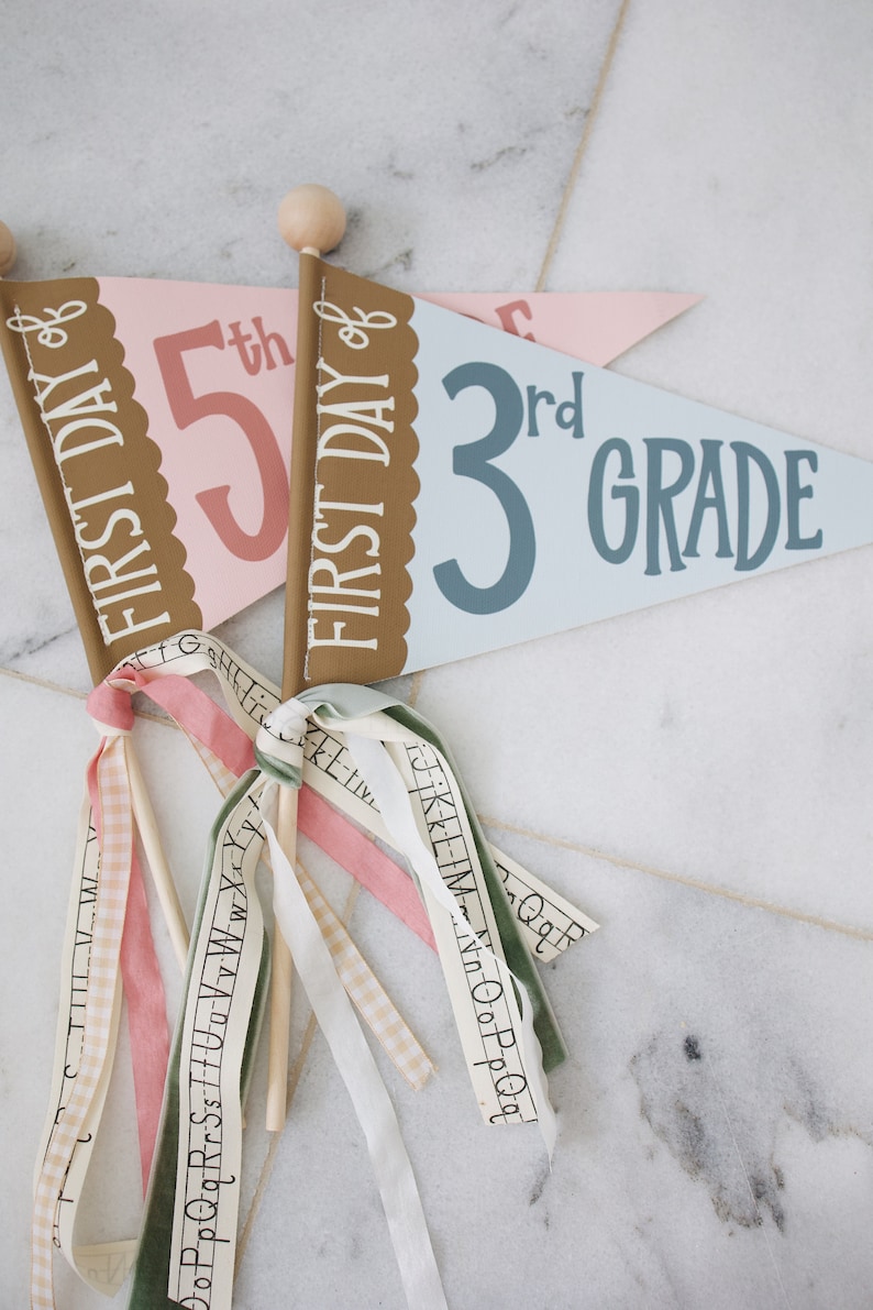 May include: Two pennant flags with wooden sticks and colorful ribbons. The first flag is pink with the text "FIRST DAY of 5th" in brown and pink. The second flag is blue with the text "FIRST DAY of 3rd GRADE" in brown and blue.