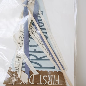 May include: A light blue and white fabric banner with the text "First Day" and a wooden stick with a blue ribbon attached. The banner is in a clear plastic bag.