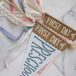 May include: Two blue and pink pennant flags with the text "FIRST DAY of PRESCHOOL" and "FIRST DAY of" on them. The flags are attached to wooden sticks with white, blue, and pink ribbons.