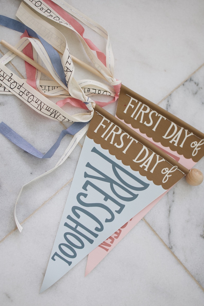 May include: Two blue and pink pennant flags with the text "FIRST DAY of PRESCHOOL" and "FIRST DAY of" on them. The flags are attached to wooden sticks with white, blue, and pink ribbons.