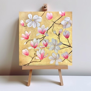 Peut inclure: Une peinture de fleurs de magnolia sur un fond doré. Les fleurs sont roses et blanches et sont disposées dans un motif lâche et fluide. La peinture est sur un chevalet en bois.