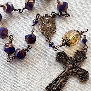 Puede incluir: Un rosario con cuentas facetadas moradas y bronce, conectadas por alambre de bronce. Presenta un crucifijo detallado y una pieza central decorativa con una imagen religiosa. Las cuentas tienen aproximadamente 8 mm de diámetro.
