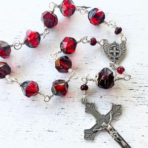 May include: A rosary with faceted, deep red beads strung on silver wire. The rosary features a silver crucifix with a detailed figure of Jesus and a decorative centerpiece. The beads are a mix of round and faceted shapes.