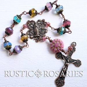 May include: A handcrafted rosary with colorful glass beads in shades of blue, purple, yellow, and pink. The rosary features a decorative butterfly centerpiece and a detailed crucifix. The text "RUSTIC ROSARIES" is visible at the bottom.