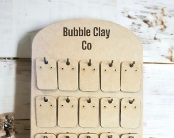May include: A wooden earring display stand with the text "Bubble Clay Co" at the top. The stand has multiple rectangular slots for displaying earrings. The stand is made of light-colored wood and has a simple, functional design.