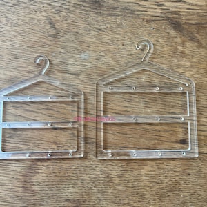 May include: Two clear acrylic hanger templates, one smaller than the other, with a hanger hook design at the top. Each template has a rectangular opening in the centre and small holes along the edges. The templates are on a wooden surface.