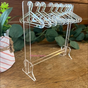 May include: Clear acrylic miniature clothing rack with matching hangers. The rack has a rectangular base and a horizontal bar for hanging clothes. The hangers are small and detailed. The text "BubbleClayCo" is visible on the rack.