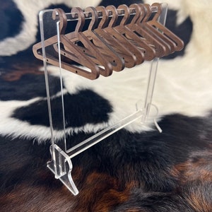 May include: A set of ten miniature wooden hangers in a dark brown colour, hanging on a clear acrylic stand. The hangers are arranged in a row, with the hooks facing forward. The stand is made of clear acrylic and has a rectangular base.
