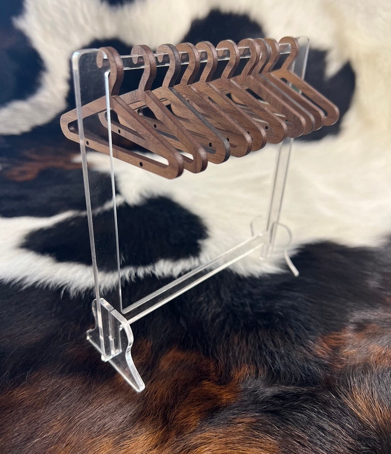 May include: A set of ten miniature wooden hangers in a dark brown colour, hanging on a clear acrylic stand. The hangers are arranged in a row, with the hooks facing forward. The stand is made of clear acrylic and has a rectangular base.