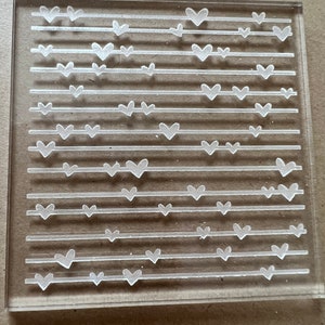 May include: Clear acrylic stamp with a repeating pattern of white hearts on horizontal lines.