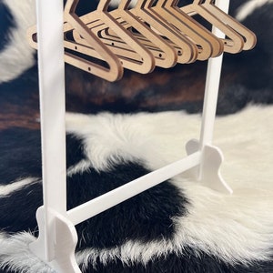 May include: A white wooden clothes rack with a horizontal bar and a set of 10 wooden hangers. The hangers are all the same size and shape, and they are arranged in a row on the bar. The rack is standing on a black and white cowhide rug.