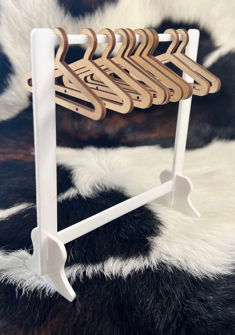 May include: A white wooden clothes rack with a horizontal bar and a set of 10 wooden hangers. The hangers are all the same size and shape, and they are arranged in a row on the bar. The rack is standing on a black and white cowhide rug.