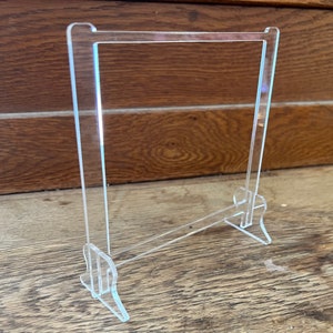 May include: A clear acrylic display stand with a rectangular base and a vertical panel. The stand is designed to hold a variety of items, such as photos, artwork, or small objects.