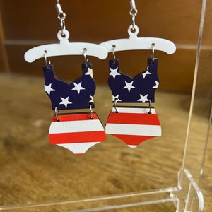 May include: A pair of wooden earrings shaped like a swimsuit with a red, white and blue American flag pattern.