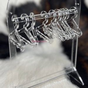 May include: A clear acrylic earring holder with a circular top and a stand. The holder has multiple hooks for hanging earrings.