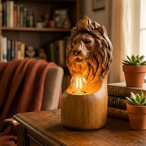 May include: A wooden lion head lamp with a warm light bulb. The lamp is brown and sits on a wooden table. The background includes books and plants. The lamp is a decorative item, and the overall aesthetic is warm and inviting.