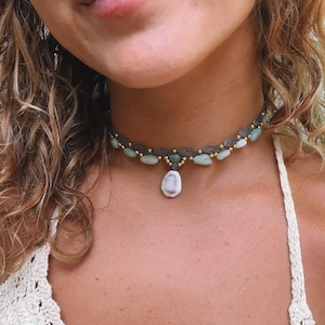 Può includere: Una collana girocollo in macramè fatta a mano con perline verde chiaro e oro. Un ciondolo bianco a forma di goccia pende al centro. La collana è indossata sulla pelle e un top color crema.