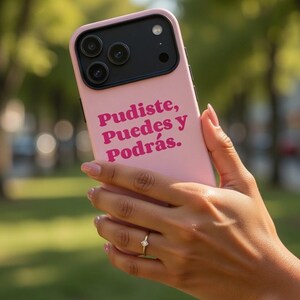 May include: A pink phone case with a black camera module. The case features the text "Pudiste, Puedes y Podrás" in a magenta font. The phone is held in a hand with a gold bracelet and ring.