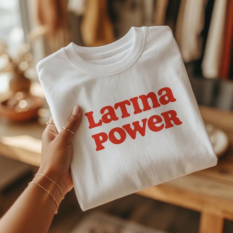 以下が含まれることがあります： 「Latina Power」の赤い文字がプリントされた白いTシャツ。 シャツは折りたたまれ、金色の指輪とブレスレットをつけた手で持たれています。 背景はぼやけており、お店の環境を示唆しています。