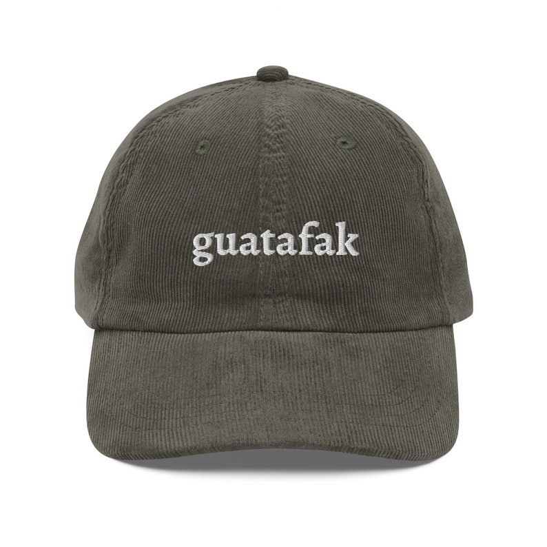 May include: Olive green corduroy baseball cap with the word "guatafak" embroidered in white. The cap has a curved brim and an adjustable strap at the back. The hat is made of a soft, textured fabric.