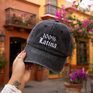 Pode incluir: Um boné de beisebol cinza-carvão com um acabamento desbotado e desgastado. A frente do boné apresenta o texto bordado em branco 100% Latina em uma fonte de estilo gótico. O boné tem uma aba curva e uma coroa estruturada de seis painéis.