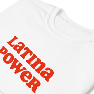 以下が含まれることがあります： 「Latina power」の文字がオレンジ色の太字でプリントされた白いTシャツ。テキストはシャツの前面に斜めに配置されています。クルーネックです。