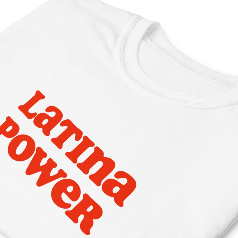 以下が含まれることがあります： 「Latina power」の文字がオレンジ色の太字でプリントされた白いTシャツ。テキストはシャツの前面に斜めに配置されています。クルーネックです。