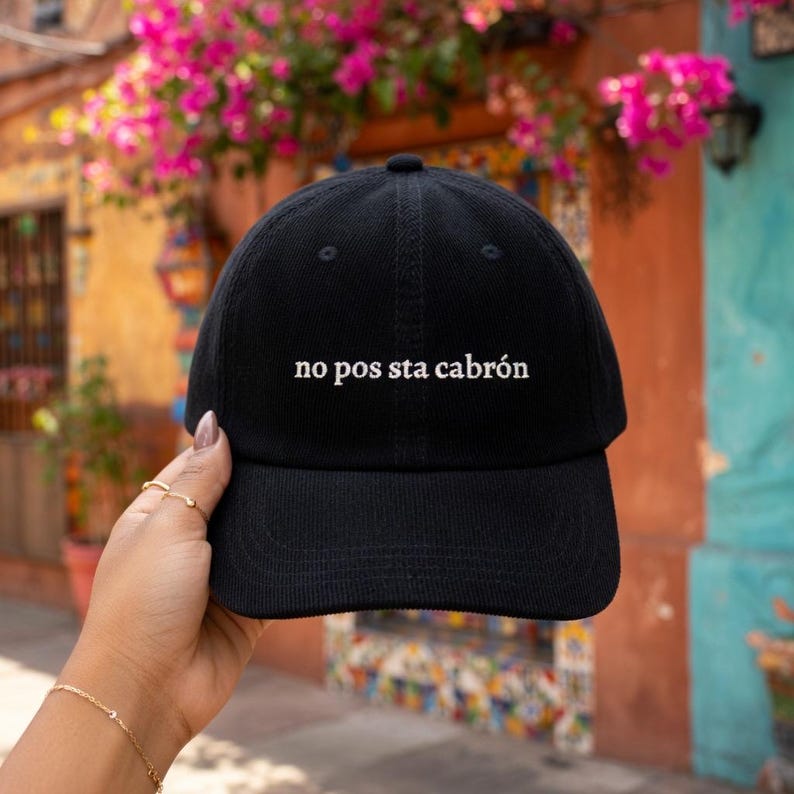 Puede incluir: Gorra de b&eacute;isbol negra con el texto "no pos sta cabr&oacute;n" en blanco. La gorra est&aacute; hecha de un material suave y tiene una visera curva. El fondo presenta edificios coloridos y flores rosas.