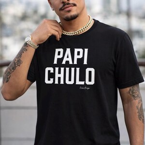 May include: Black t-shirt featuring the words "PAPI CHULO" in large white capital letters. Underneath, the phrase "Eres fuego" is printed in a smaller font. The shirt has a crew neck design.
