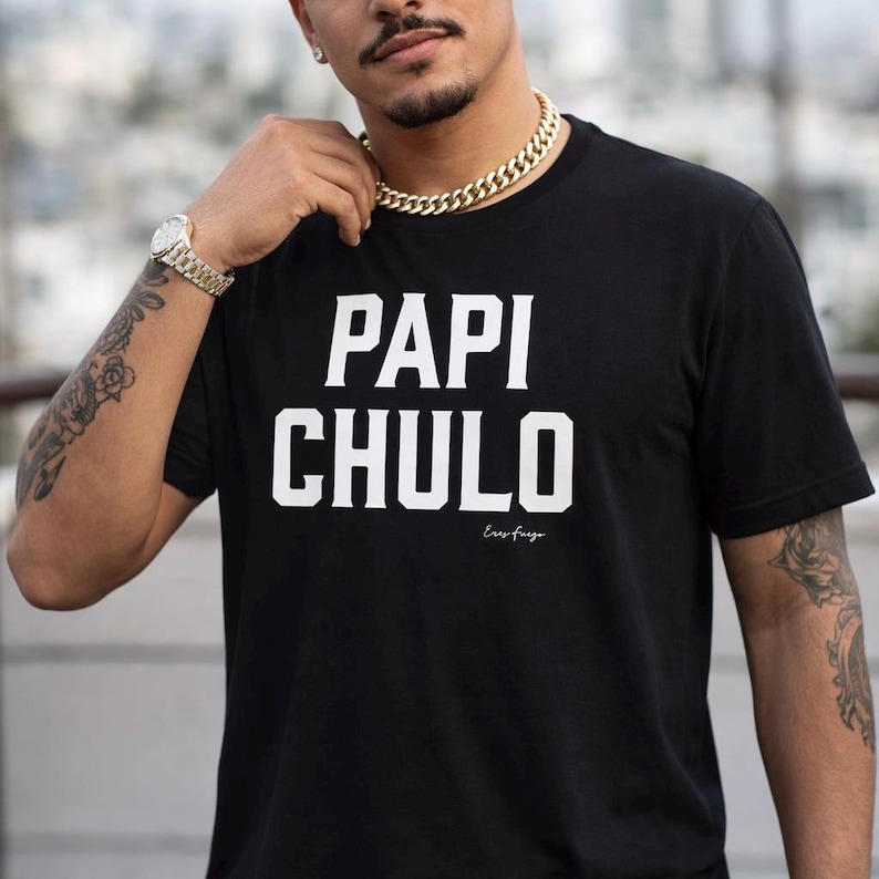 May include: Black t-shirt featuring the words "PAPI CHULO" in large white capital letters. Underneath, the phrase "Eres fuego" is printed in a smaller font. The shirt has a crew neck design.