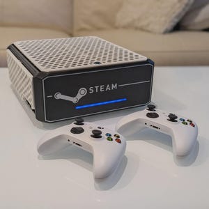 May include: A black and white Steam console with a perforated top and the word STEAM printed on the front. Two white game controllers with black buttons and colourful accents are in front of the console.