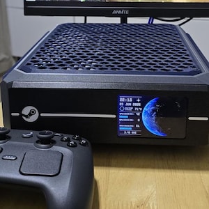 May include: A black gaming computer console with a honeycomb-patterned top vent and a front-facing digital display showing system statistics, including CPU temperature of 71 degrees Celsius, alongside a black wireless game controller.