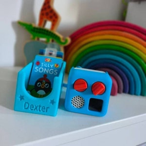 May include: A light blue plastic music player holder with the name Dexter embossed on the front, containing a small book titled Silly Songs with a cartoon bird graphic. Next to it is a matching blue speaker unit with two red knobs and a speaker grille.