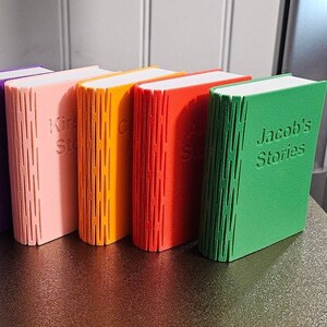 Selection of 3d printed storage boxes for holding Yoto cards. They are designed to look like a book, have a colour cover, white pages, and can be customised with any text.