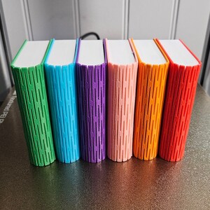 Selection of 3d printed storage boxes for holding Yoto cards. They are designed to look like a book, have a colour cover, white pages, and can be customised with any text.