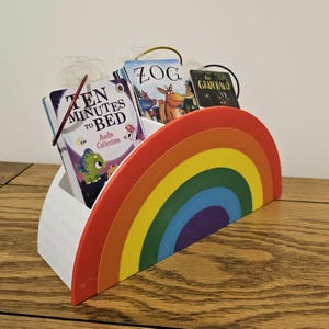 May include: A rainbow-shaped book holder made of white plastic with a red, orange, yellow, green, blue, indigo, and violet colour scheme. The holder is filled with three children's books: "Ten Minutes to Bed", "Zog", and "The Gruffalo".