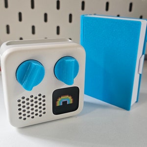 May include: A white electronic device with two blue knobs and a rainbow pixelated graphic. The device is next to a blue book.