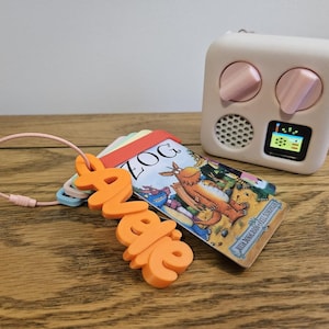 May include: A light pink audio player with two knobs, a speaker grille, and a small digital screen, positioned next to a set of plastic cards on a pink wire ring. One orange plastic tag features the name "Avelle" in raised, bold, sans-serif letters.